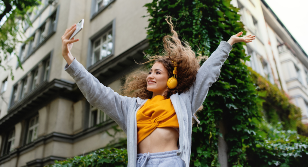 Young woman with great skin and curly hair wearing yellow headphones, taking a selfie outdoors for Tik Tok in an urban setting with greenery.