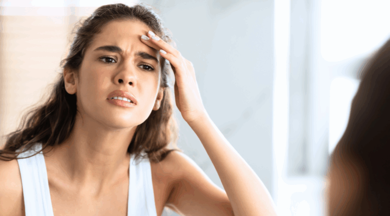 Young woman examining her forehead for wrinkles in the mirror, questioning if it's the right time to start anti-wrinkle injections
